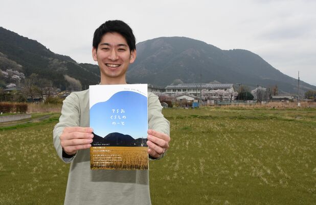 寺尾地区で親しまれるゾウが寝そべったような形の三峰山を背に、冊子をＰＲする国府谷さん
