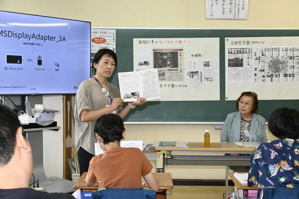小金井空襲体験者の簗さん（右奥）から話を聞いた後、副読本に掲載された小金井空襲のページを示しながら児童に語りかける稲見教諭＝１３日午前、下野市細谷小
