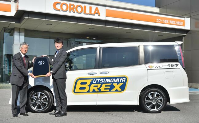 寄贈された乗用車の前で喜谷社長（左）から鍵のレプリカを受け取る藤本社長＝宇都宮市上横田町