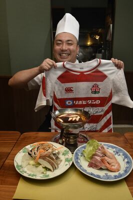 選手らのサイン入りユニホームを手にする石田さん。手前の料理はイタリア遠征で提供した料理の火鍋＝６日午後、鹿沼市内