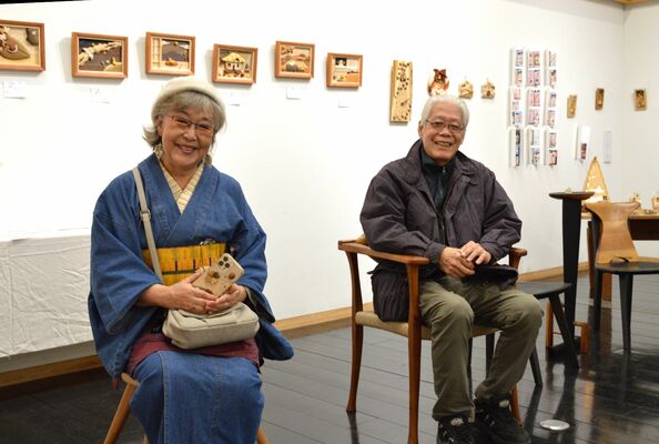 展示会場で大島さん（左）の額絵作品を背景に、神田さん（右）の作品の椅子に座る２人