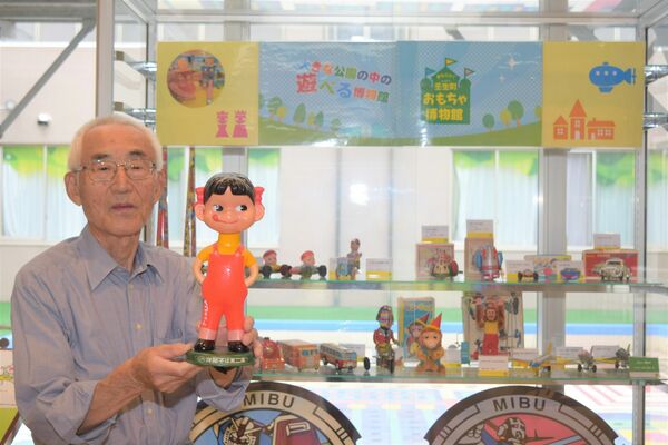 寄贈したおもちゃを背に、「ぺこちゃん首振り人形」を手に持つ粂川さん