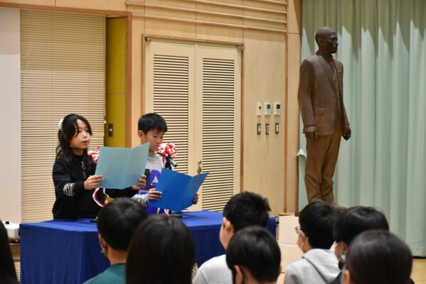 お披露目された田村の立像を脇に、創立130周年記念集会のあいさつをする代表児童