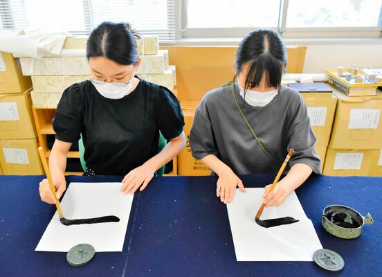 愛媛大大学院生の河野亜美さん（右）が左手で、岡田優佳さんが右手での毛筆を実演。横画を書くとき、右手では筆を引き、左手では筆を押す動きになる＝７月末、松山市文京町の愛媛大