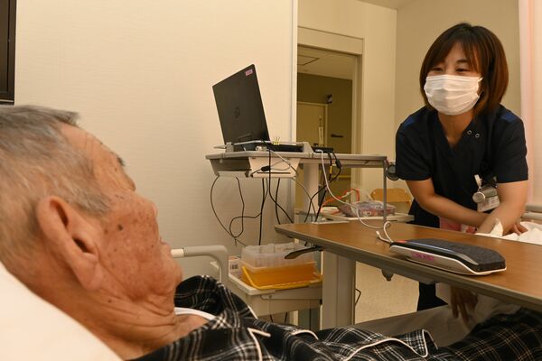 患者に容体を尋ねる看護師（右）。会話はマイク機器（右手前）を通じて自動で文字に起こされる＝とちぎメディカルセンターしもつが