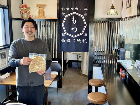 真空パックのモツ煮を掲げる山崎さん