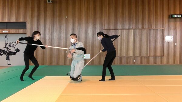 動画内で女子社員2人に拘束される佐野社長(中央)