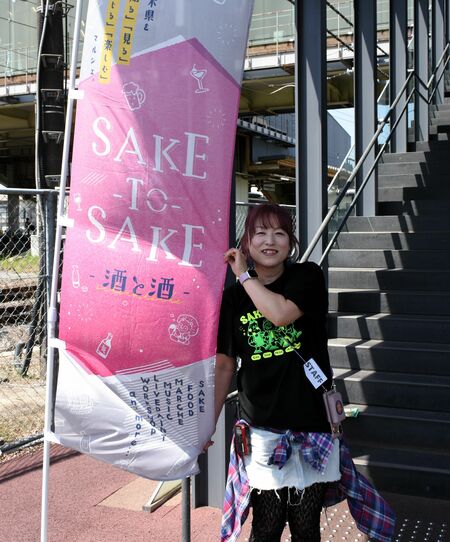 「ＳＡＫＥ＿ＴＯ＿ＳＡＫＥ」のぼり旗に手を添える星絵理子さん（右）＝４月２６日、ＪＲ宝積寺駅前